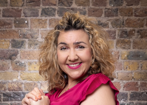 Amy J Payne &ndash; mezzo-soprano, actor and director &ndash; smiling in a professional portrait, wearing a vibrant pink top against an exposed brick background.