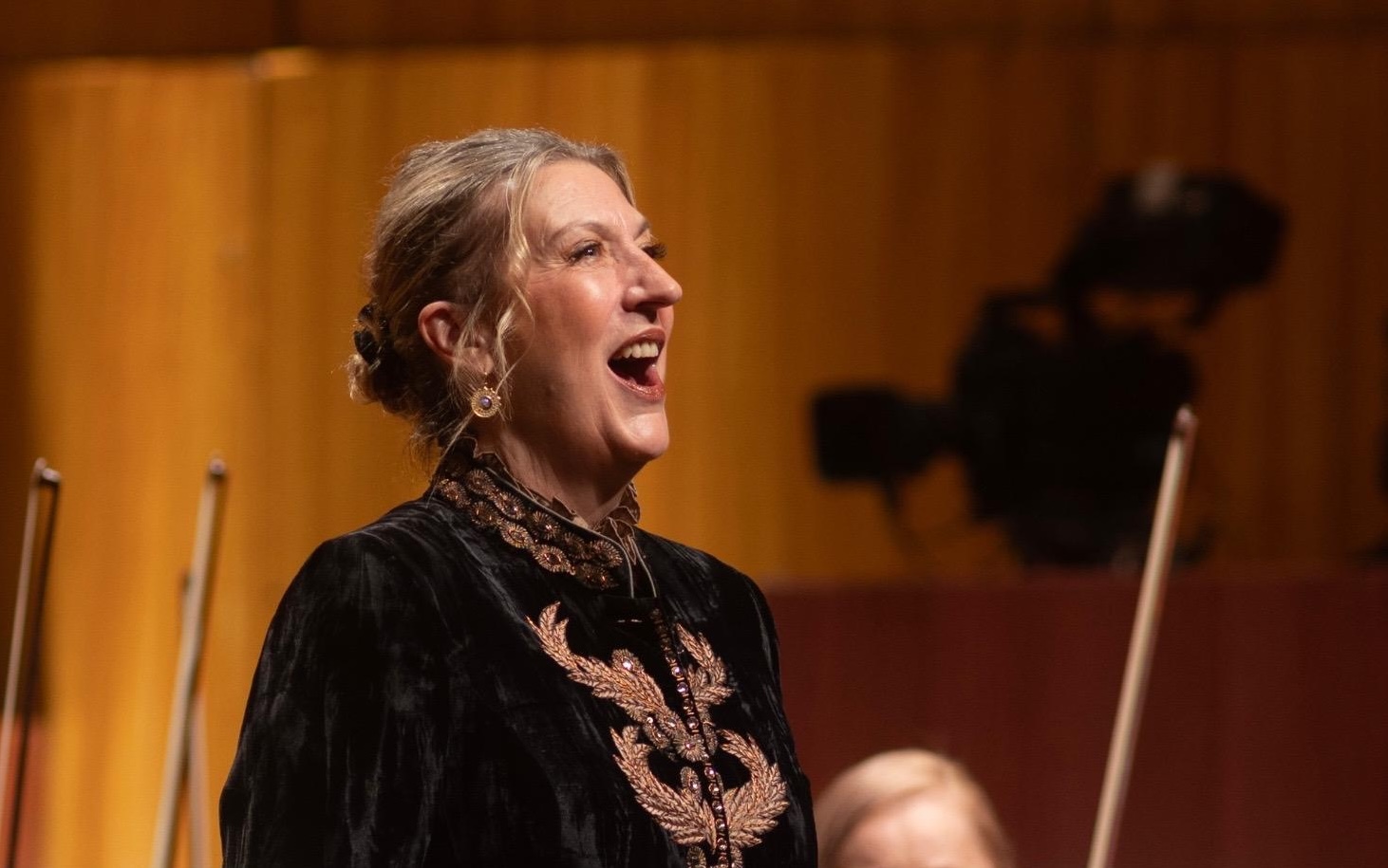 Dame Sarah Connolly mid-performance, singing on stage in formal black concert attire with gold detailing, warmly lit against an orchestral backdrop.