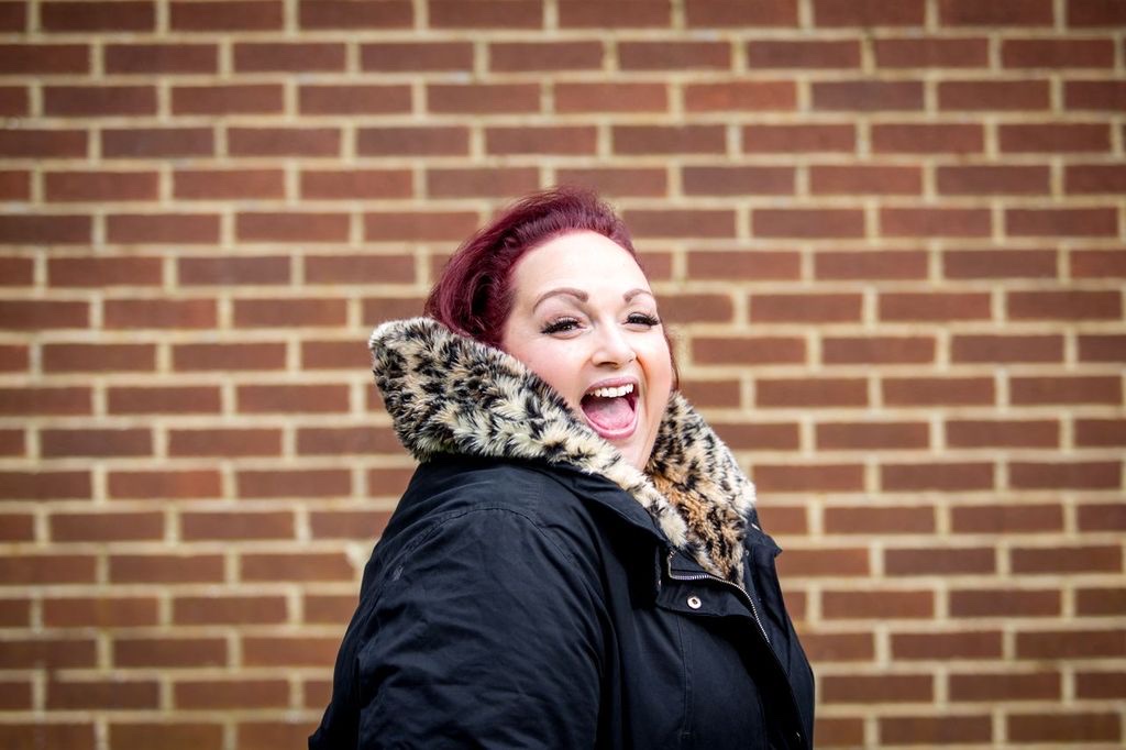 Helena Dix, soprano, laughing in a black coat with a faux-fur collar against a brick wall.