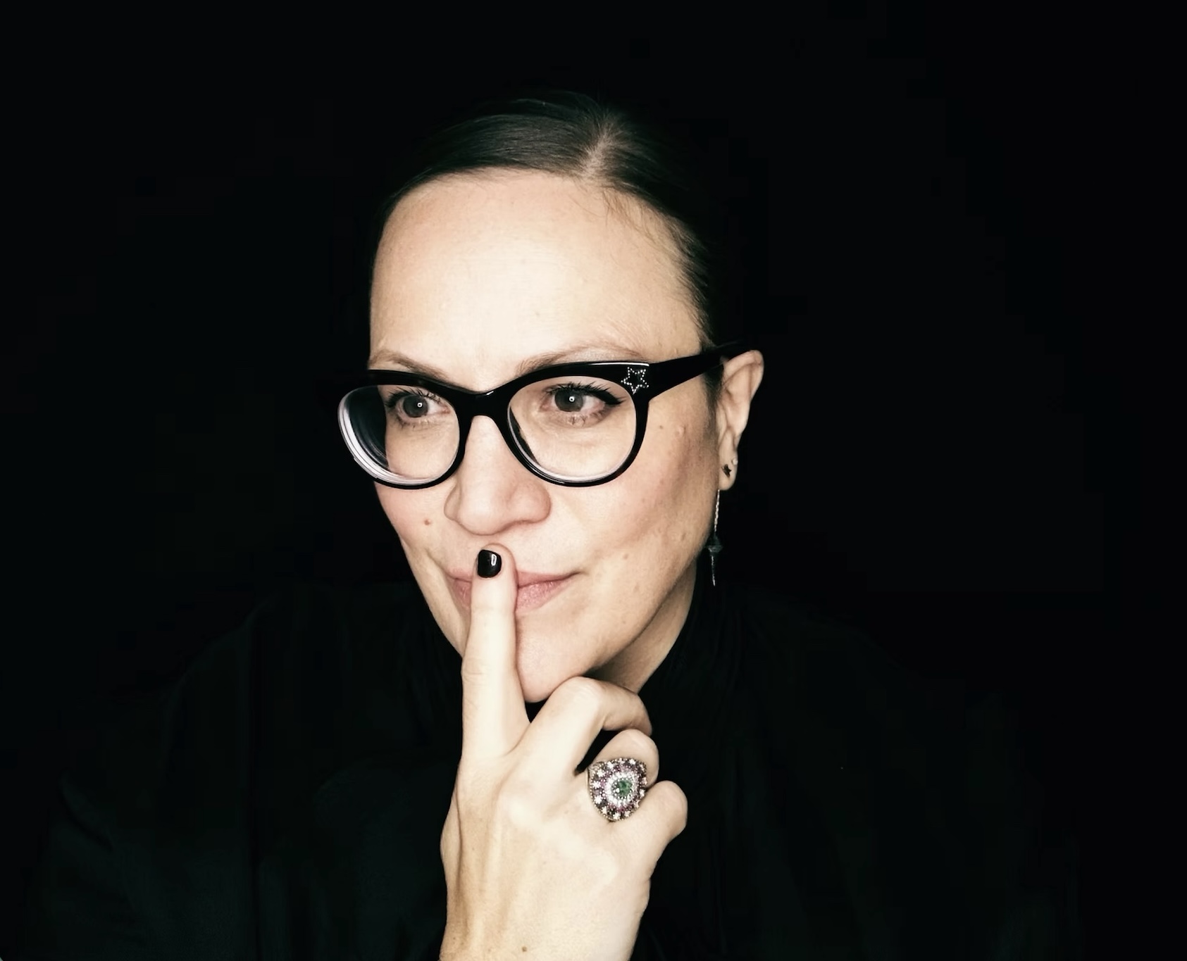 Music Makers: with Jennifer Johnston, mezzo-soprano A striking portrait of mezzo-soprano Jennifer Johnston against a deep black background. Dressed entirely in black, with her dark hair pulled neatly back, she is captured in a moment of quiet reflection. Wearing distinctive black cat-eye glasses, she gazes thoughtfully to one side, her finger resting lightly against her lips in a contemplative pose. Subtle jewellery, including an ornate statement ring, adds detail to the otherwise minimalist composition. Soft, focused lighting highlights her expression and creates an intimate, dramatic atmosphere.