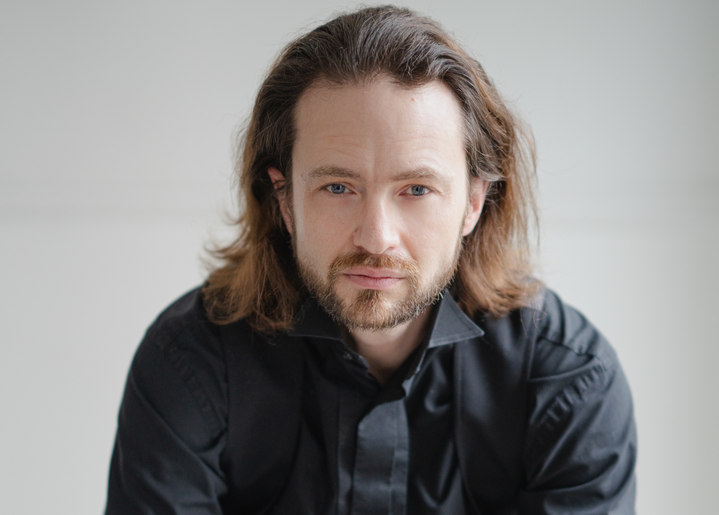 Portrait of conductor John Andrews, looking directly at the camera, wearing a black shirt against a neutral background.
