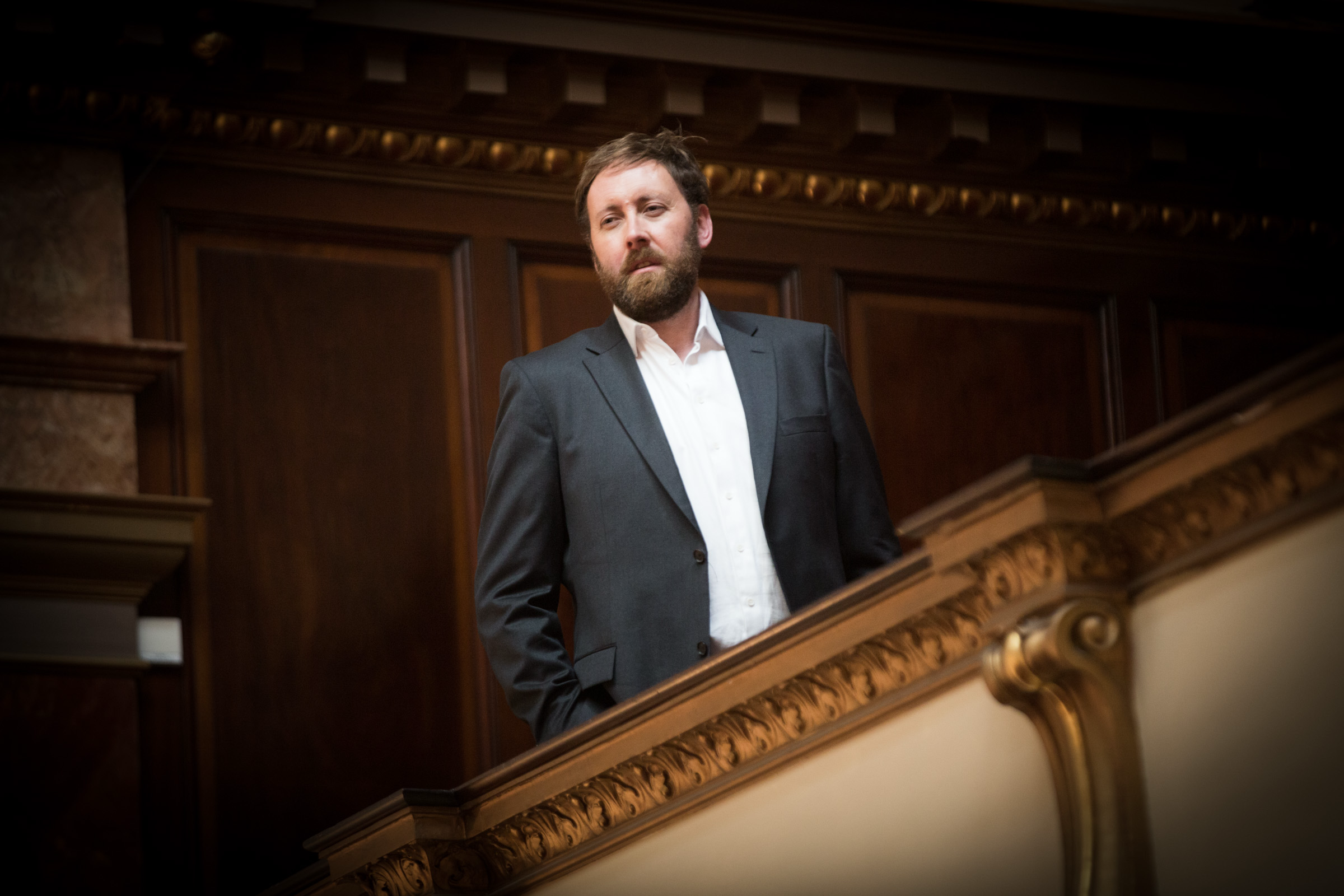 Matthew Best - Bass Bass Matthew Best stands on an ornate staircase in a concert hall, looking into the distance, wearing a grey suit jacket and open-collar white shirt.