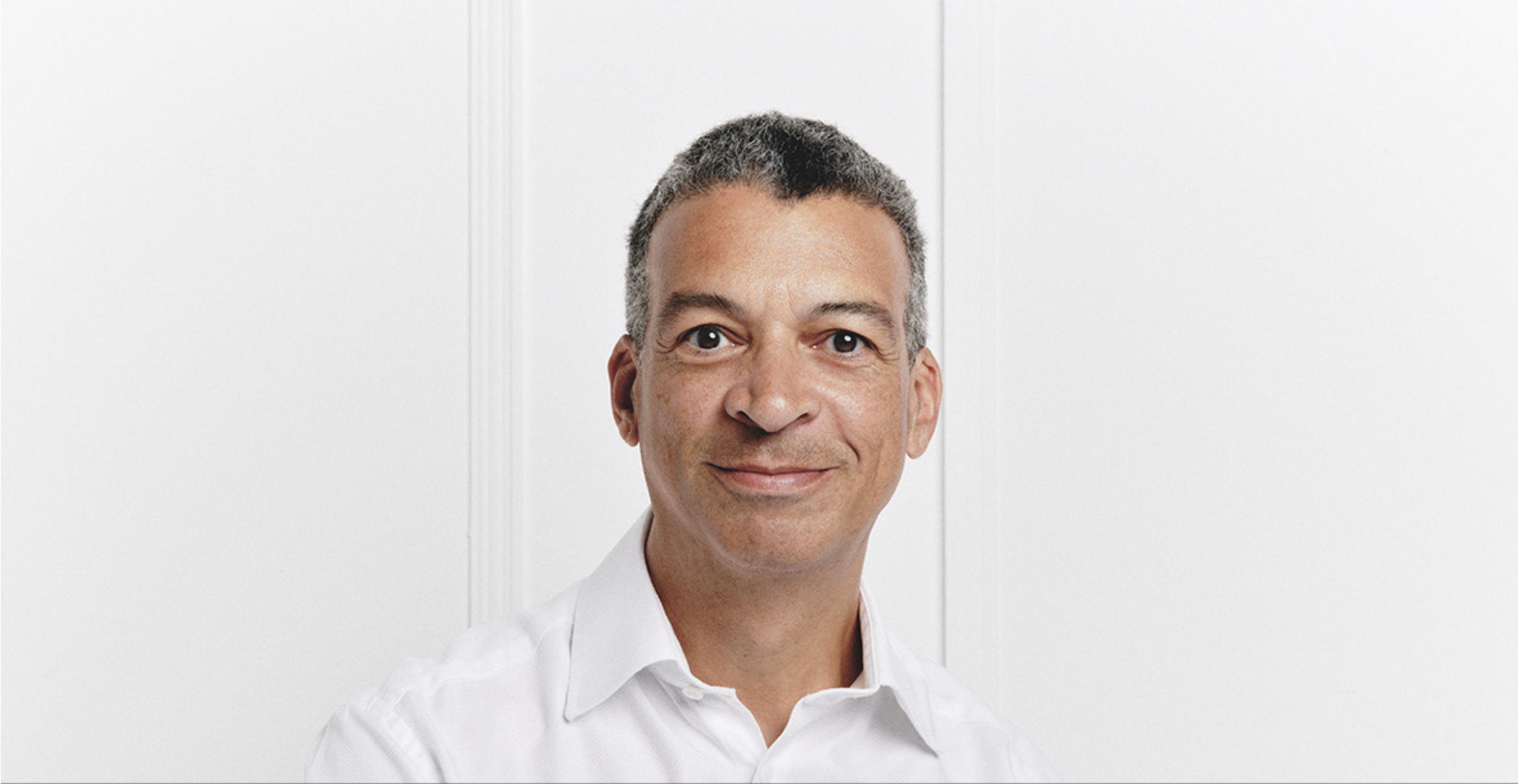 Portrait of Roderick Williams, wearing a white open-collar shirt, facing the camera with a calm expression against a soft grey background.
